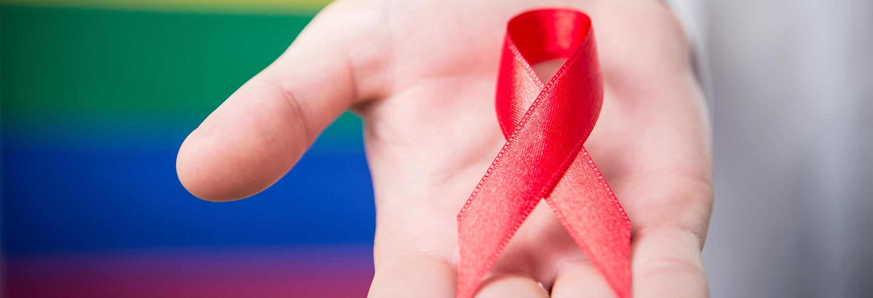 close up image of HIV/AIDS red ribbon on a palm of a hand in front of a rainbow wall